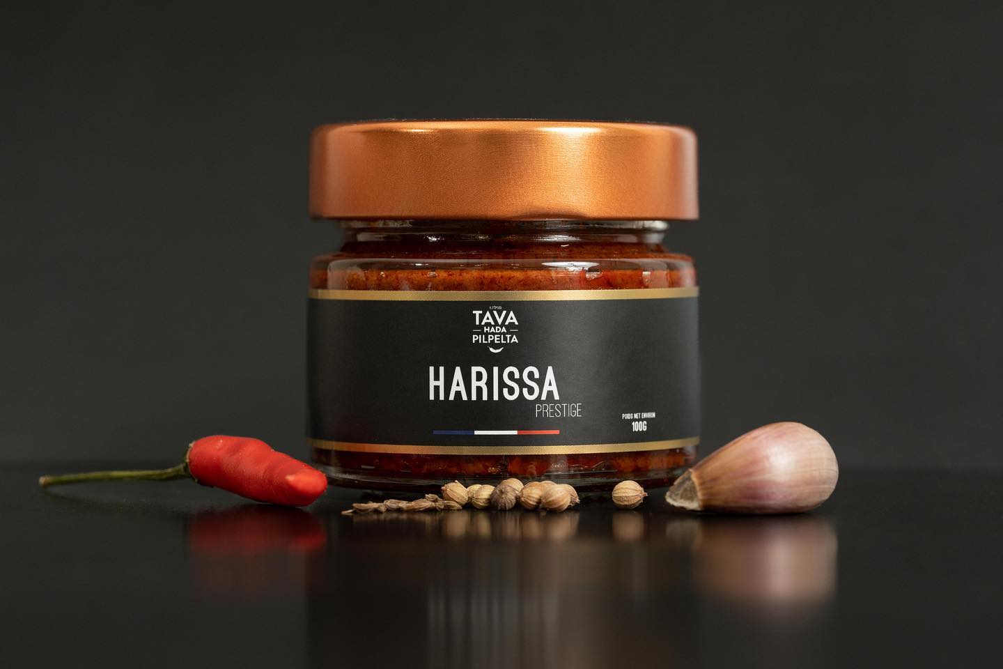Pot de harissa prestige Tava Hada Pilpelta sur fond sombre avec un piment et une gousse d'ail