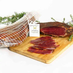 Jambon ou viande séchée tranchée sur planche de bois, décorée de herbes et filet de protection sur le côté gauche.